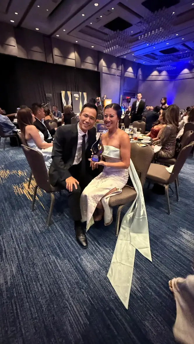 Maxine Sudarma seated at a gala table holding the AACC Community Impact Award