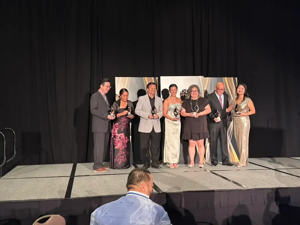 Seven AACC 2026 award honorees holding their awards on stage, with Maxine Sudarma at center